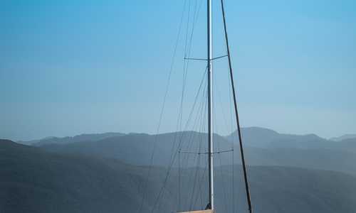 A sailboat rests comfortably in the morning sun. The sailboat accompanied by green hills in the background, as the city seems distant and small. The clear blue skies above, the sun hitting poles where sails would hang, while below it, calmed rippling water reflecting its beautiful brown hull back at itself.